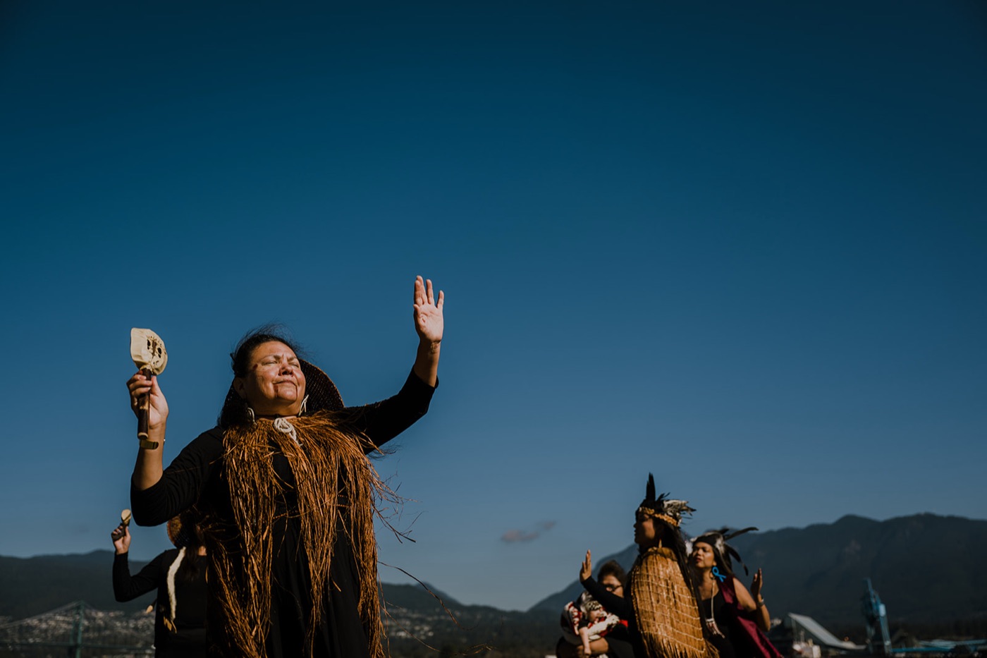 A woman in a cedar-bark cape dances with a rattle raised, others dancing behind her beneath the mountains.